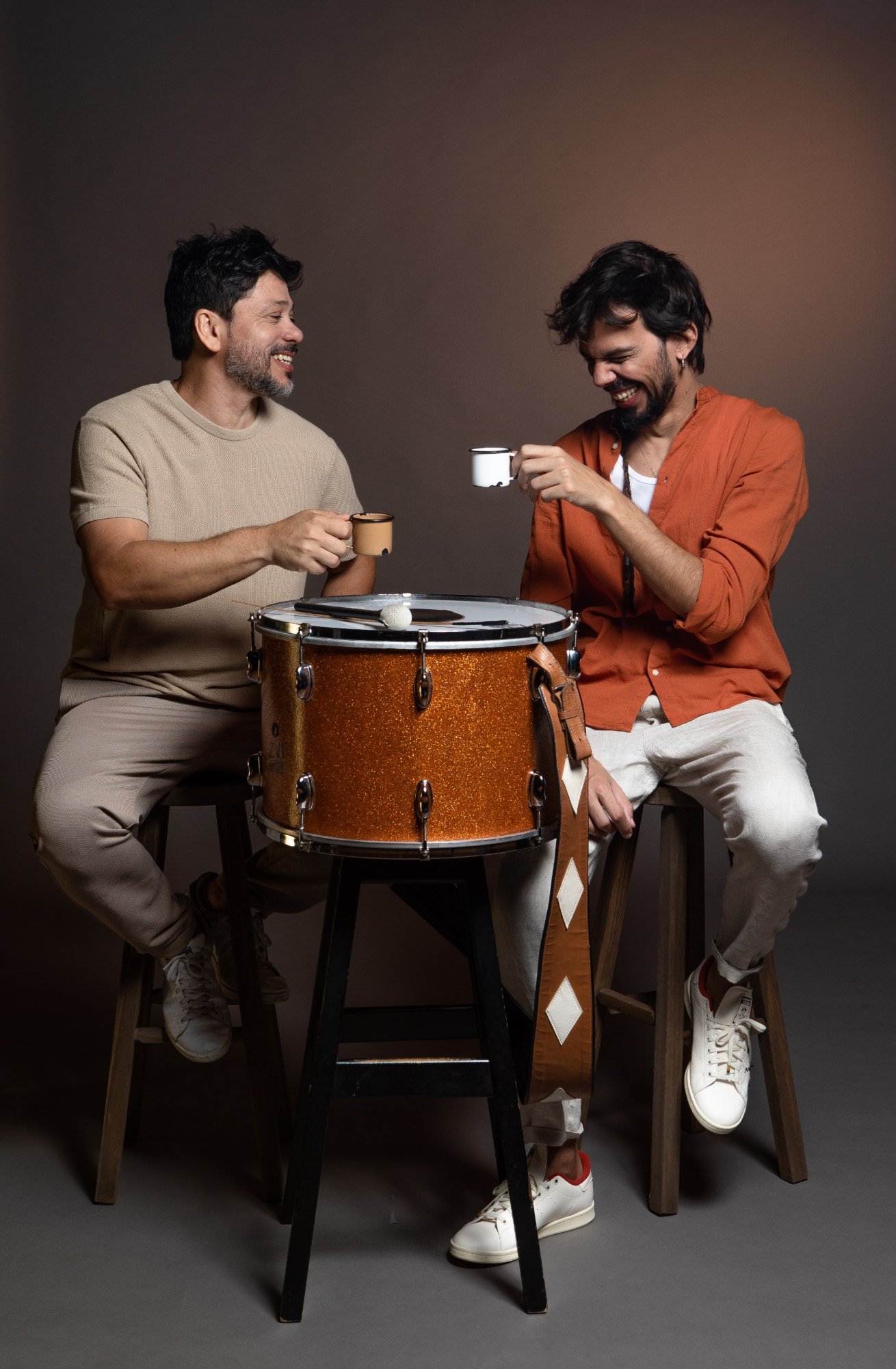 Guegué Medeiros and Salomão Soares laughing and toasting with coffee mugs, sitting around a zabumba