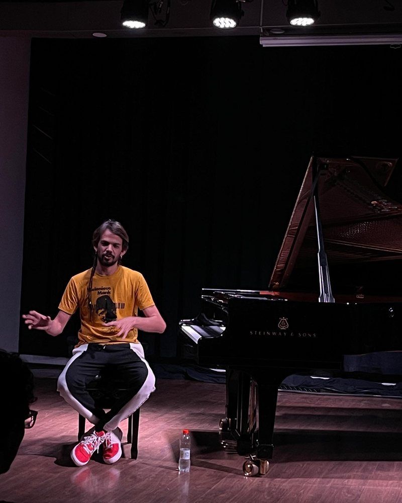 Salomão Soares falando ao público, sentado ao lado do piano de cauda
